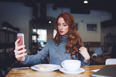 Boğazlı kazaklı, sakin, zarif bir kadın. Modern kafede bir fincan sıcak içecek ve pastayla kahvaltı ederken akıllı telefondan selfie çekiyor.