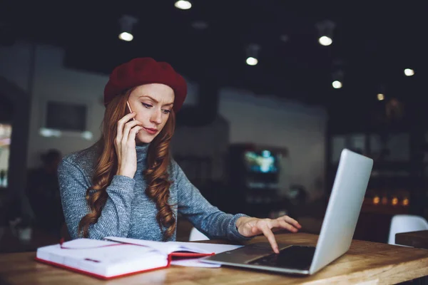 年轻而专注的女性远程工作者坐在桌旁 一边在笔记本电脑上打字 一边用手机聊天 一边在现代的工作空间里独自工作 — 图库照片