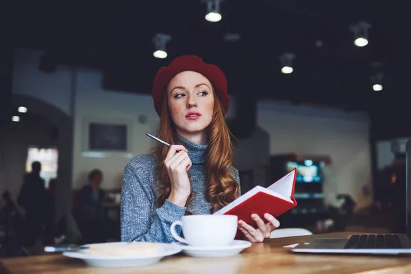 在早餐的时候 穿着红色贝雷帽 沉默寡言 优雅的红头发女人一边在咖啡店的红皮笔记本上写字 一边若有所思地回头看 — 图库照片