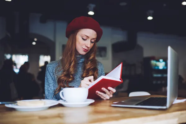 穿着高领毛衣 头戴红色贝雷帽的长发女人专心致志地在笔记本上用红色封皮写字 同时在咖啡店远程操作笔记本电脑 — 图库照片