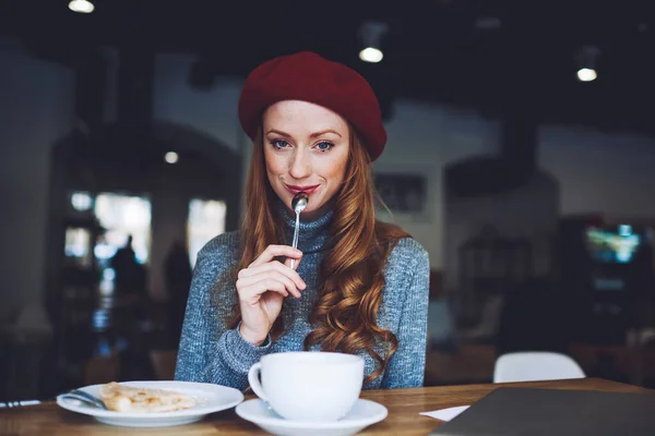 若いです幸せな長い髪の女性で赤ベレー帽飲む熱い飲み物と持っているデザートでカメラで居心地の良いカフェテリア — ストック写真