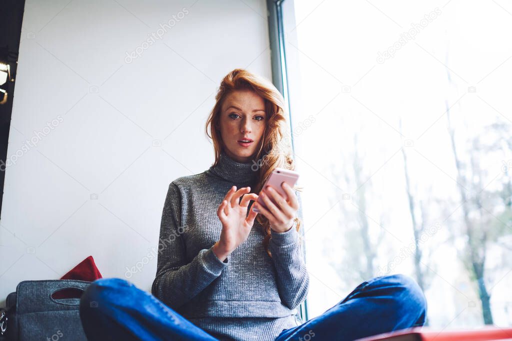 Low angle of serious female in casual clothes looking at camera sitting on windowsill with crossed legs chatting with friends having free time