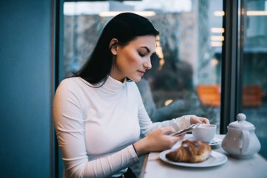 Uzun siyah saçlı, kruvasan ve kahve içen cep telefonlu genç bir kadının yan görüntüsü.