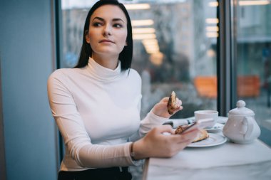 Genç konsantre kadın kruvasanla masada oturuyor ve kafede mola sırasında sosyal ağlarda mesajları kontrol etmek için akıllı telefon kullanıyor.