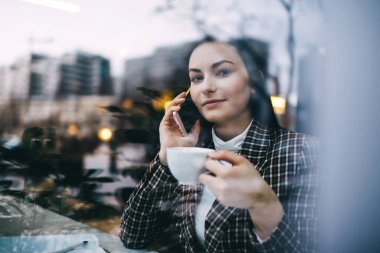 Kendine güvenen, şık giyinmiş, kahve fincanında kameraya bakan ve akıllı telefondan konuşan uzaktan kumandalı bir kadın.