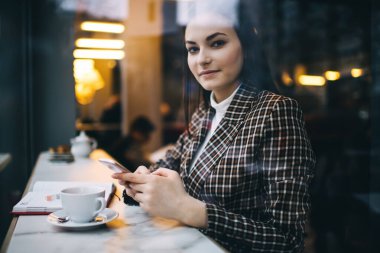 Moda kıyafetli, elinde kahve fincanı ve cep telefonu ile masaya oturmuş kameraya bakan mutlu kadın uzaktan işçinin penceresinden.