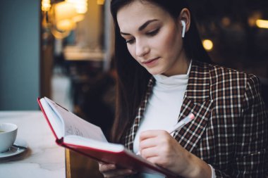 Zarif kıyafetli ve kulaklıklı genç bir kız öğrenci masada oturup modern kafede müzik dinlerken not defterine not alıyor.