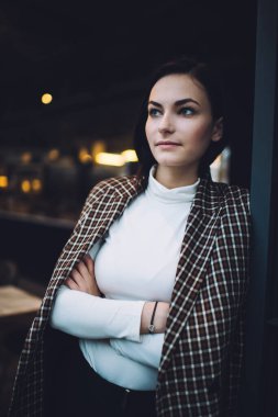 Modern kafede kollarını kavuşturmuş duvara yaslanmış siyah saçlı, kendine güvenen genç bir kadın başka tarafa bakıyor.