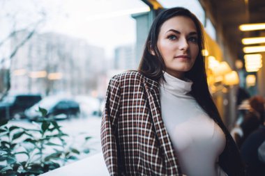 Uzun siyah saçlı, ekose ceketli, çağdaş bir kafede ahşap masanın yanında duran ve gün ışığında başka tarafa bakan olumlu genç bayan.