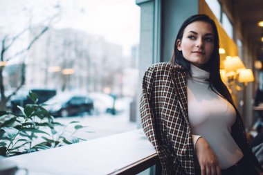 Zarif ceket giyen düşünceli genç bir bayan ve modern ışıklı kafede masaya yaslanmış beyaz balıkçı yaka kazak giyip gündüz vakti başka tarafa bakıyor.