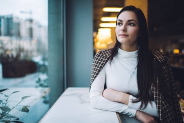 Uzun siyah saçlı, beyaz balıkçı yakalı, beyaz ellerle beyaz masaya yaslanmış ve kafeteryada boş vakit geçiren düşünceli genç bayan.