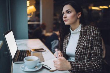 Genç serbest çalışan, şık kıyafetli bir bayan kahve ve dizüstü bilgisayarla masada oturuyor ve kafede kağıda not alıyor.