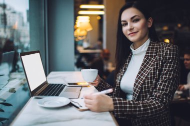 Genç gülümseyen kız öğrenci sıcak içecek içerken test için hazırlanıyor kafeteryada dizüstü bilgisayarla oturuyor ve kameraya bakıyor.