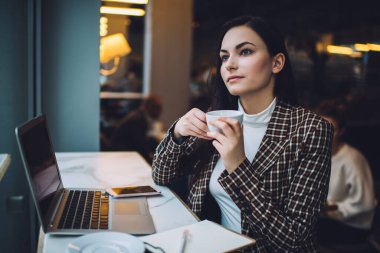 Genç, düşünceli, serbest çalışan, şık giyinen, sıcak bir şeyler içen, kafede dizüstü bilgisayarlı bir masada oturan genç bir bayan.