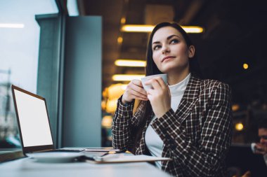 Düşünceli genç bayan iş kadınının düşük açılı görüntüsü şık günlük kıyafetler giyip kahve içerken ve kafeteryada dizüstü bilgisayarla otururken başka yere bakarken.