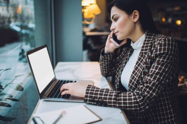 Ciddi bir bayan yöneticinin ofiste çalışırken cep telefonuyla konuşma ve dizüstü bilgisayarla yazı yazma görüntüsü