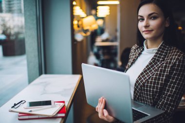 Zarif kıyafetler içindeki genç bir kadın kafeteryada elinde not defteri ve akıllı telefonuyla masanın yanında durup kameraya bakarken uzaktan bilgisayarla çalışıyor.