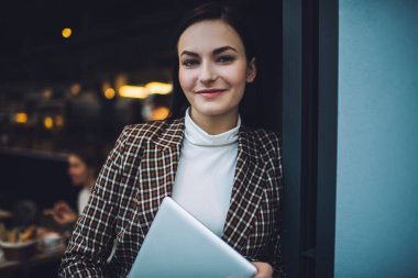 Ellerinde netbook ile gün içinde kafeteryanın girişine yaslanıp kameraya bakarken zarif giysiler içinde gülümseyen genç bayan.