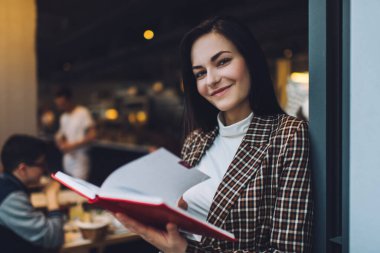 Kahvede defterin sayfalarını çevirirken siyah uzun saçlı, kareli ceketli ve beyaz bluzlu gülümseyen genç bir kadın.