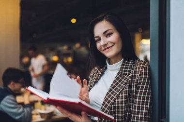 Gün içinde kafeterya duvarına yaslanıp kameraya bakarken elinde açık bir defterle, zarif giysiler içinde gülümseyen genç bir kadın.