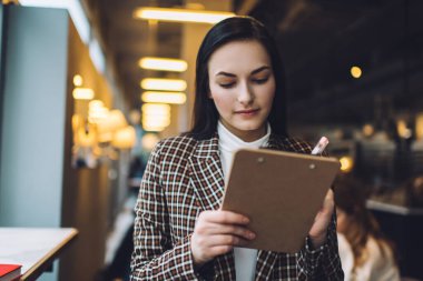 Kareli ceketli, beyaz bluzlu, siyah, uzun saçlı, kalemle kafedeki panoya yazı yazan odaklanmış genç bir kadın.