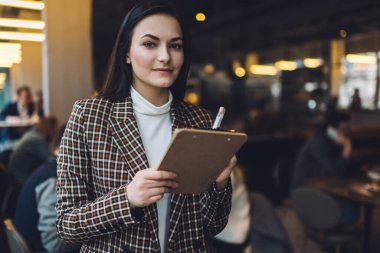 Kareli ceketli ve beyaz bluzlu genç bir kadın kafedeki panoya kağıt yazarken kameraya bakıyor.