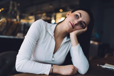 Genç, sakin, beyaz elbiseli bir kadın kafeteryada oturuyor, başını eğiyor ve elini uzatıyor.