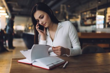 Genç ve ciddi içerikli bir kadın cep telefonuyla konuşuyor ve kafenin bulanık arka planındaki masada defter inceliyor.