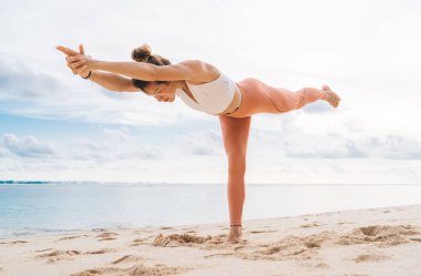 Denize yakın kumsalda yoga yaparken Warrior III Asana 'da çıplak ayaklı, çıplak ayaklı genç bayanın düşük açılı görüntüsü.