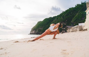 Aktif giyimde çıplak ayaklı fit bir kadın vücudu tropikal tatil beldesinde kumlu kıyılarda okyanus kıyısındaki eğitim sırasında Baddha Parsvakonasana yoga pozu veriyor.