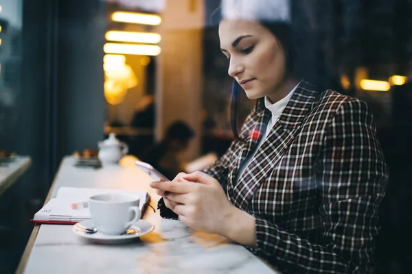 カフェのテーブル席に座ってコーヒーを飲みながらスマホでメッセージを発信するスタイリッシュな服を着た女性の集中的な側面図 — ストック写真