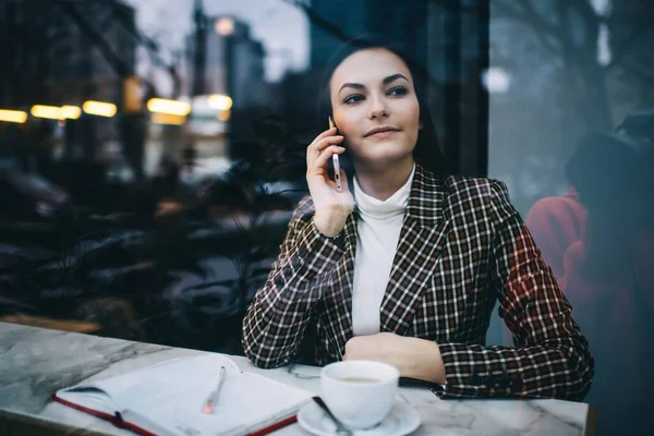 コーヒーとノートブックを持ってテーブルに座っている間に見ている幸せな女性のフリーランサーの窓を通して電話をかける — ストック写真
