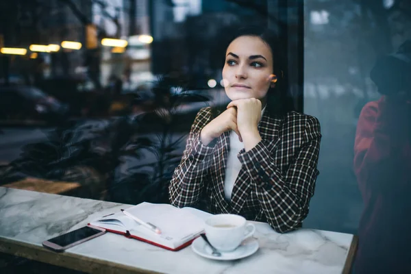 穿着时髦夹克 头脑清醒的女人坐在餐桌旁 手里拿着笔记本和一杯饮料 穿过玻璃窗 白天在咖啡店里望着远方 — 图库照片