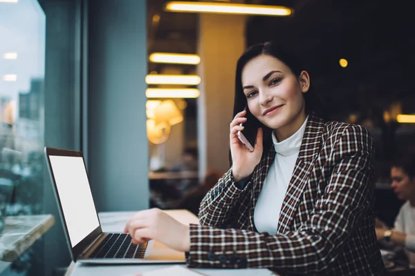 Zarif kıyafetler içindeki genç bayan bilgisayarla uzaktan yakından ilgilenirken akıllı telefondan konuşuyor ve kafeteryada oturuyor ve kameraya bakıyor.