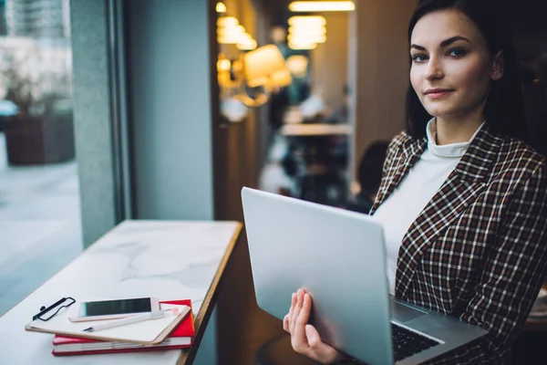 Zarif kıyafetli genç bir kadın dizüstü bilgisayarla çalışırken gün içinde kafede telefon ve not defterinin yanında durup kameraya bakıyor.