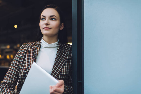 Confident young woman in elegant outfit leaning on wall while standing with computer in hands in cafe entrance in day and looking away