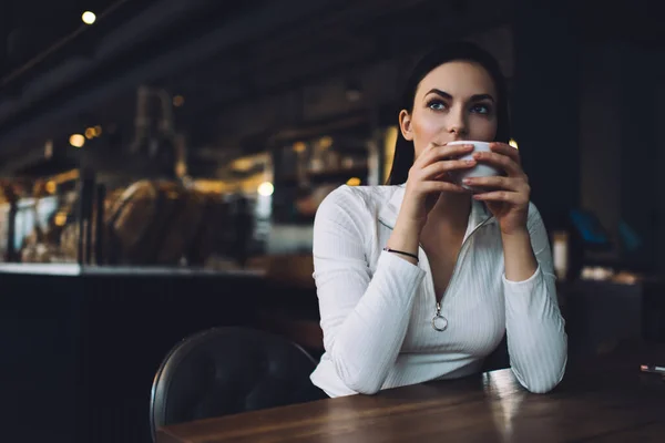 多愁善感的年轻女性 深色长发 白衬衫 喝着瓷杯里的热饮料 坐在咖啡店旁 望着远方 — 图库照片