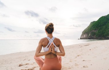 Güneşlenirken ve meditasyon sırasında özgürlüğün tadını çıkarırken kumlu sahilde oturan sportif bayanın arkası.