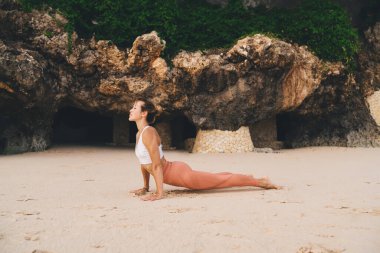 Aktif kıyafetli ince bir kadın yeşil kayalıkların yakınındaki kumlu sahilde Yukarı Yüzlü Köpek Asana 'ya bakıyor ve sağlıklı bir hayat sürerken gözlerini kaçırıyor.