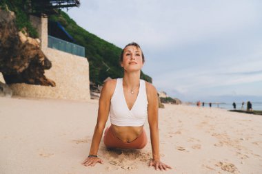 Mutlu, uyumlu bir kadın yazın bulutlu, kasvetli gökyüzünün altında kumlu sahilde köpek pozu veriyor.