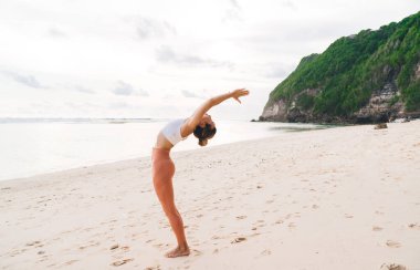 Urdhva Hastasana 'da kumlu sahilde kayalık kayalıklarla kaplı okyanus kenarındaki yeşil çalılarla genç bir kadının yan görüntüsü.