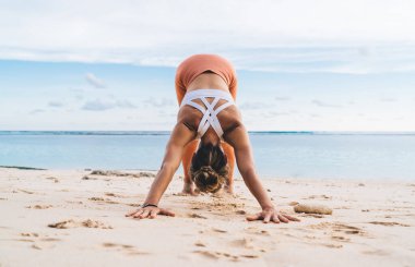 Avuçlarını kuma dayamış, kumlu havada kumsalda deniz kenarında serinlerken yoga ve eğilme egzersizleri yapan zayıf bir kadın vücudu.