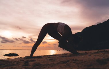 Güneş batarken renkli gökyüzünün altında sakin okyanusun kıyısındaki kumlu kumsalda yoga yapan isimsiz bir kadının silueti.