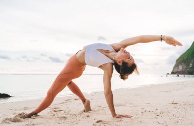 Spor giyimli çıplak ayaklı bir kadın ve kayalık plajda yoga yaparken Düşmüş Üçgen duruşunda duran bilezik takan bir kolye.