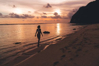Sıska bir kadının sakin okyanusun yakınındaki kumlu sahilde yürüdüğünü ve Bali 'de tatil yaparken sakin turuncu günbatımının tadını çıkardığını görüyorum.