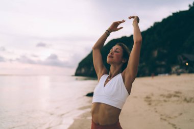 Genç zayıf sporcu kadın, bulutlu bir günde Bali 'de deniz kenarında meditasyon yaparken kollarını kaldırıp nefes alıyor.
