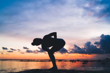 İsimsiz bir kadının silueti yoga yapıyor ve yaz mevsiminde günbatımının keyfini çıkarırken deniz kıyısında duruyor.