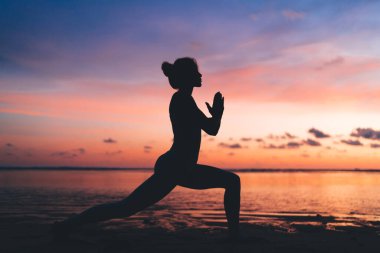 Kadın siluetinin yandan görünüşü, deniz kıyısındaki bilinç meditasyonu sırasında renkli gökyüzü ile dua etmeye devam ediyor. Namaste 'de akşam vakti dinlenen zayıf bir kadın kıyı şeridindeki fiziksel dinlenme sırasında dinleniyor.