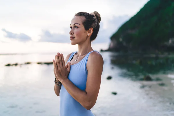 海の水と山に対して野外で瞑想しながら手のヤシを一緒に入れて海辺に立って積極的に大人の女性の側面図 — ストック写真