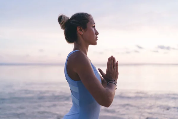 日の出のワークアウト中に海のビーチで折り畳まれた手でヨガを瞑想し 練習体のスーツで穏やかなアジアのスポーツ女性の側面図 — ストック写真